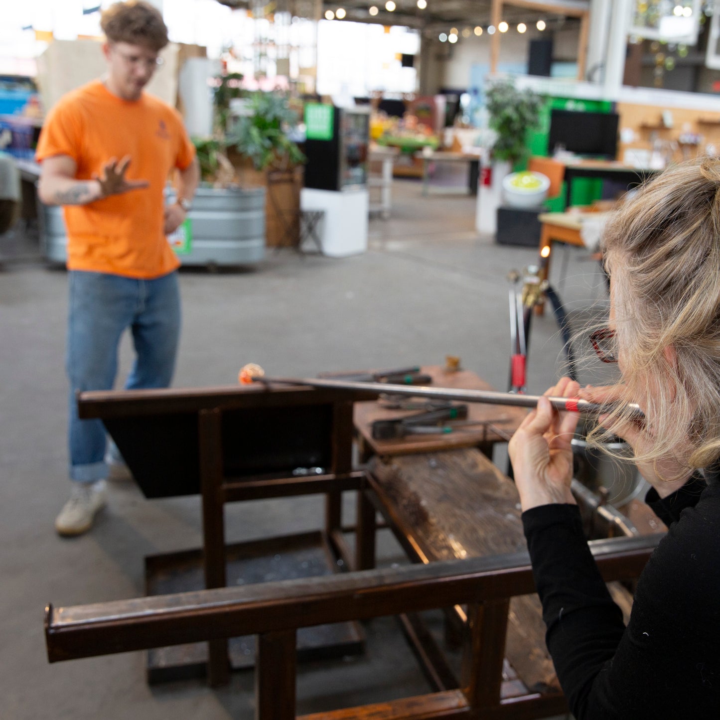 Our glassblower, Jake, instructs a 100% Hands-On class student while she uses the blowing pipe to blow a gather of molten glass into a bubble.