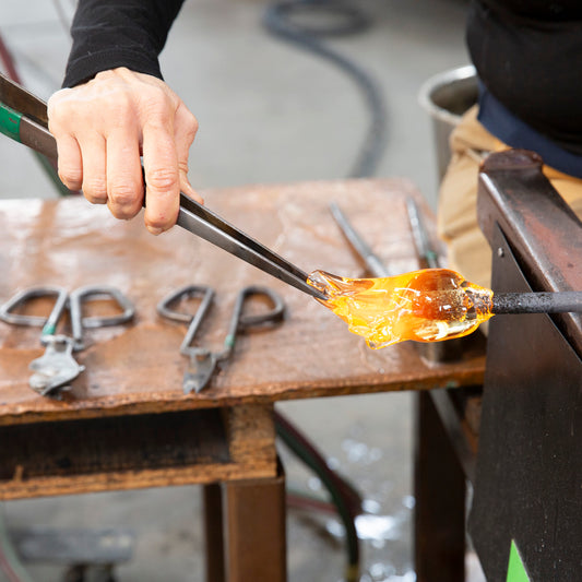 A 100% Hands-On class student works with large tweezers to pull a gather of molten glass.