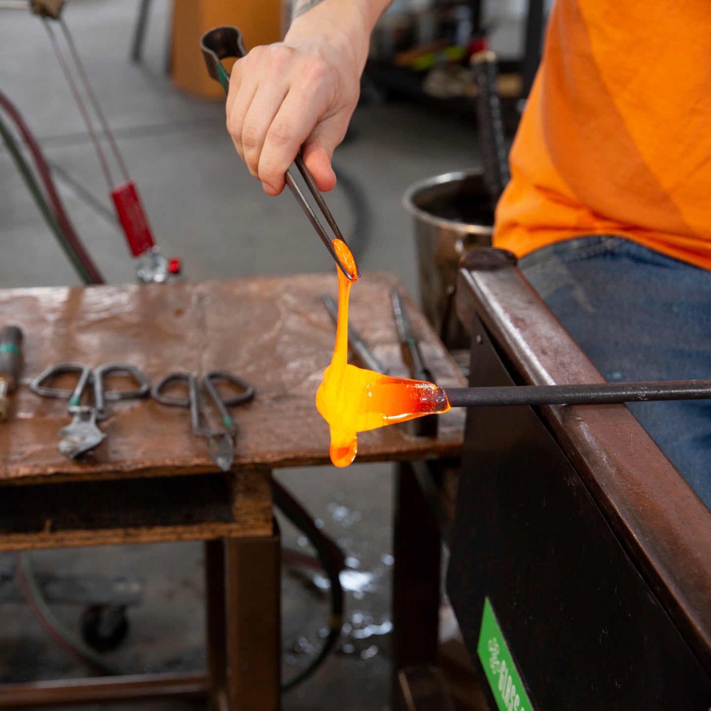 A 100% Hands-On class student works with large tweezers to pull a dripping gather of molten glass.