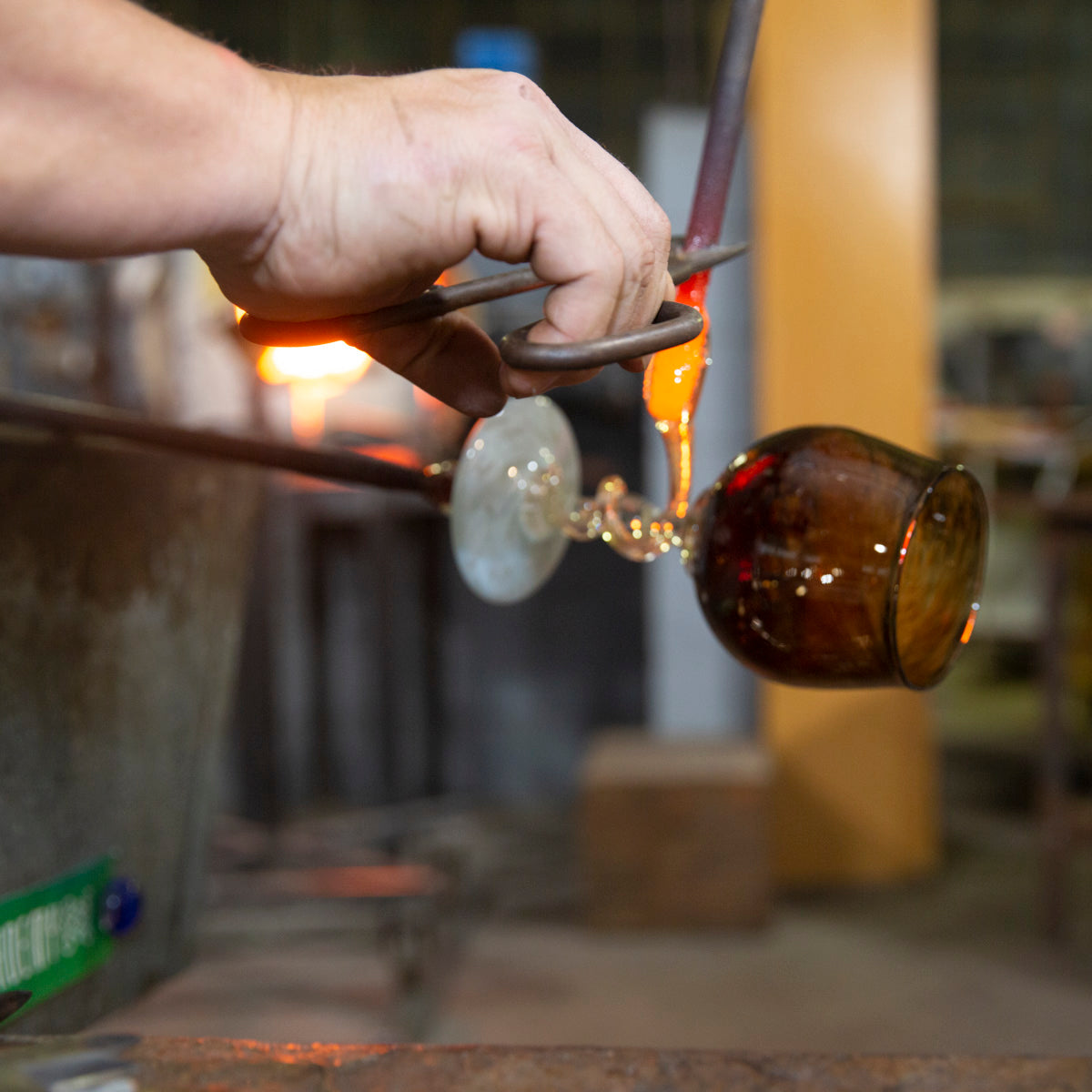 In a close-up shot, a glassblower works with molten glass to add a swirl to the stem of a Happy Hour class participant's custom wine glass.