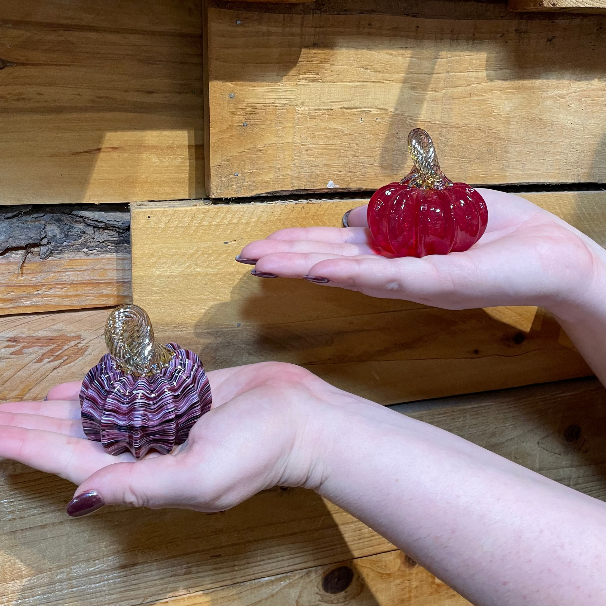 Two glass pumpkins, one red and one purple, held in hands against a wooden background.