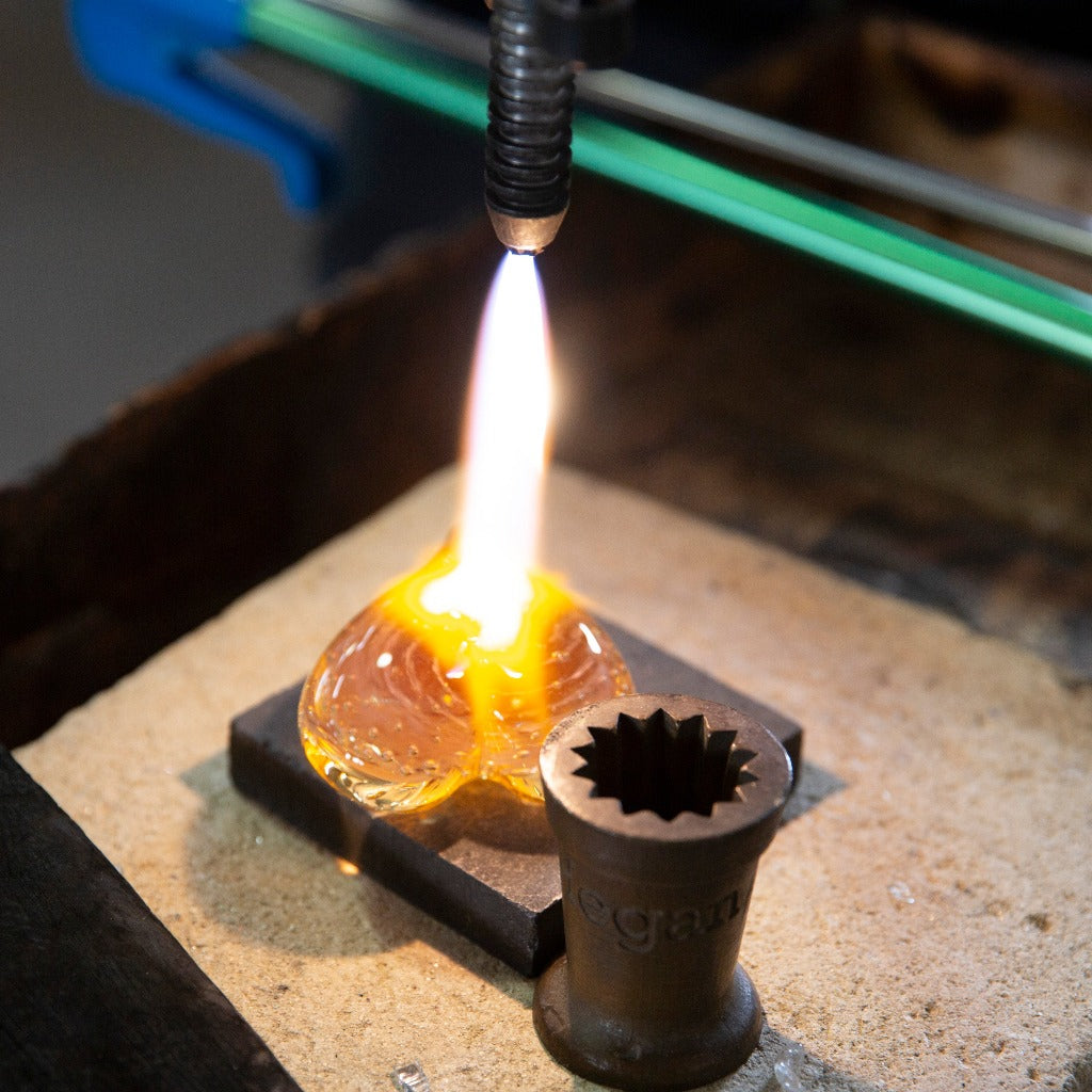 A molten glass heart is fired with a blowtorch on a piece of metal.