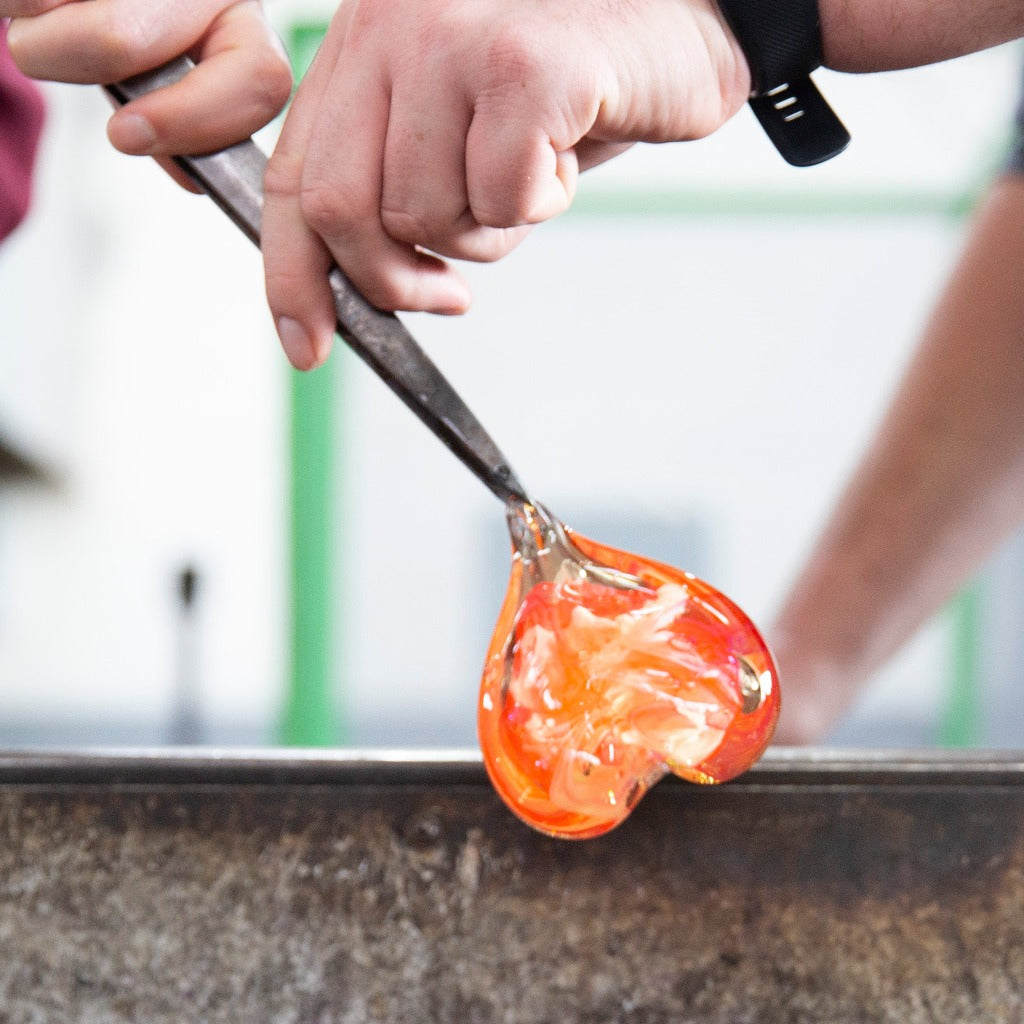 A glassblower helps a Glass Heart class student shape the bottom point of their heart with tweezers.